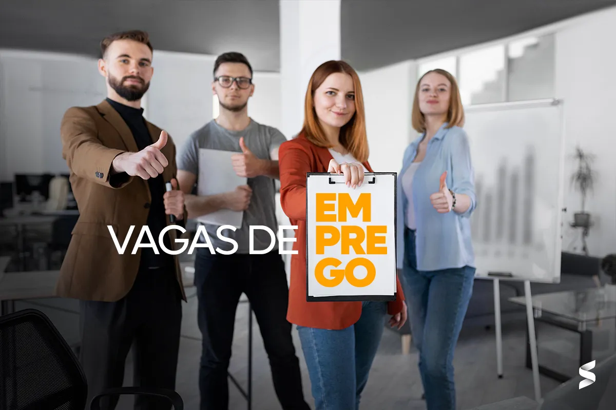 Equipe sorridente em ambiente corporativo segurando um cartaz com "Emprego".